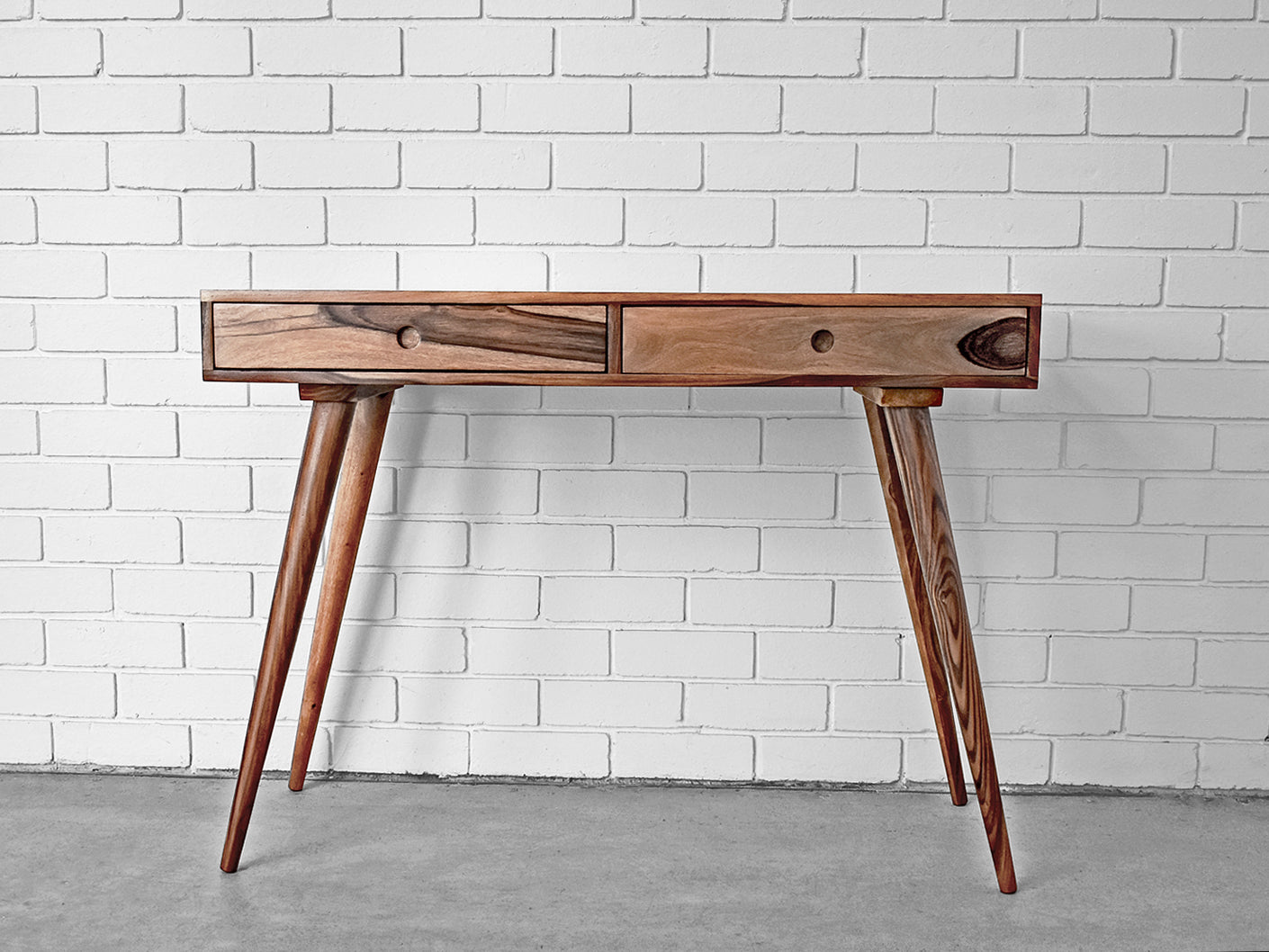 A mid-century modern wooden console table with two drawers, angled splayed legs, and a white brick wall background.
